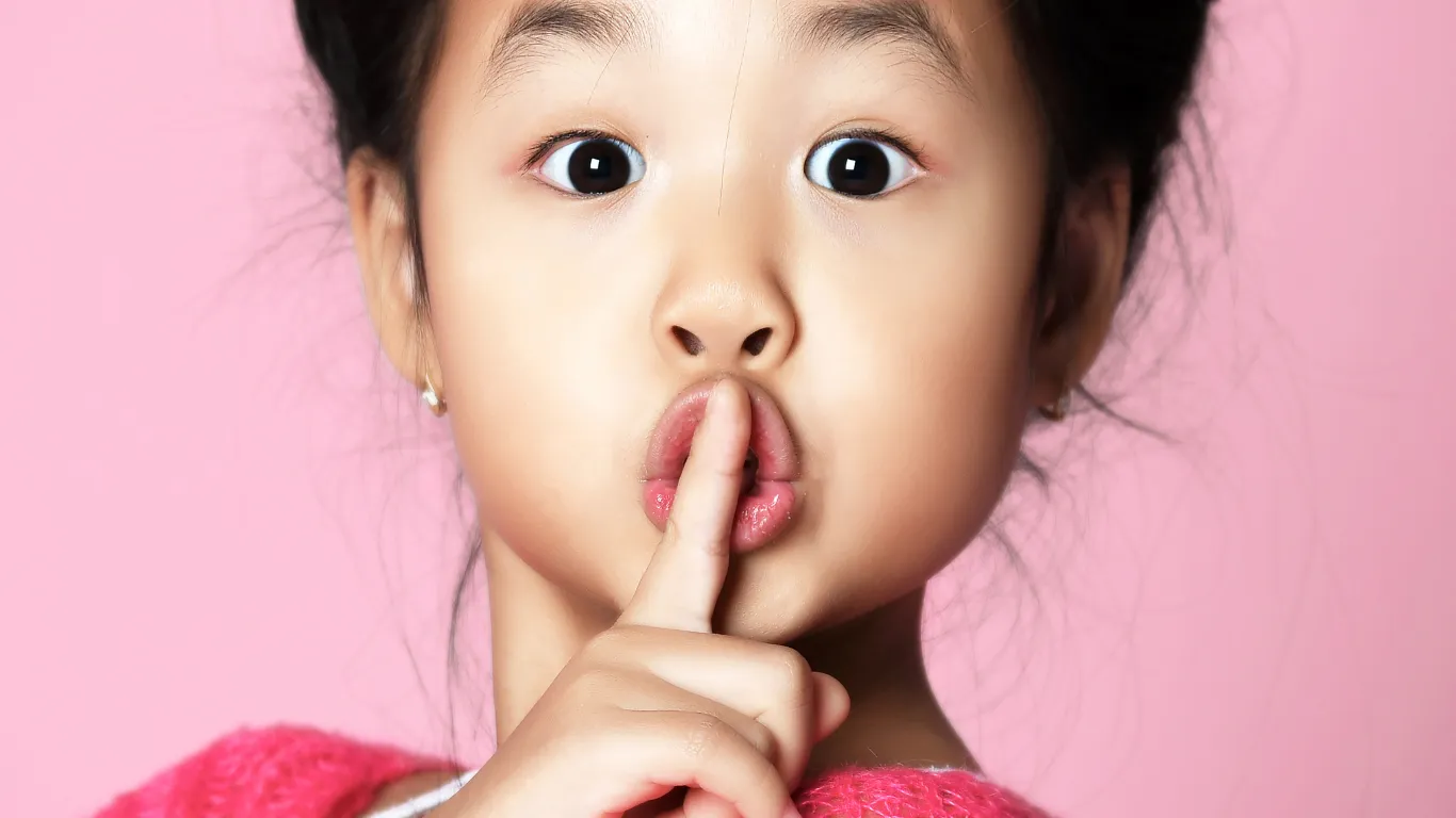 A young girl with her index finger to her lips as if urging someone to be quiet.